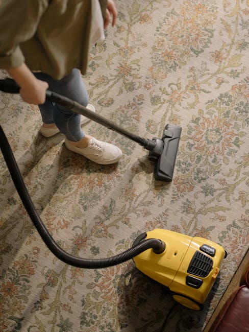 A person vacuuming a decorative area rug with a patterned floral design in a room. The individual is wearing beige sneakers, dark jeans, and a beige jacket, operating a yellow and black carpet cleaning vacuum cleaner with a long hose and metal wand attachment. The room has warm ambient lighting, and the carpet appears clean and well-maintained. The image illustrates domestic surface cleaning, emphasizing deep cleaning and sanitisation as part of professional services provided by Carpet Cleaning Harringay, aligned with their Green Lanes carpet cleaning guide for Harringay flats.