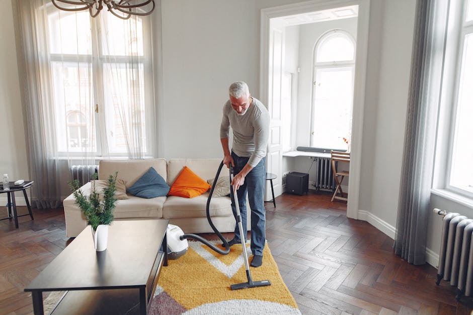 A middle-aged man engaged in domestic cleaning within a spacious, well-lit living room. He is using a handheld vacuum cleaner to perform surface cleaning on a multicolored geometric-patterned rug situated in front of a white sofa adorned with orange and blue cushions. The room features large windows with sheer curtains allowing natural light to fill the space, highlighting the warm wooden flooring arranged in herringbone pattern. Nearby, a small side table with a potted plant and a window seat with a bench are visible, alongside a radiator beneath one window and an electronic keyboard in the corner. The scene emphasizes thorough cleaning and maintenance of living room surfaces, showcasing the professional services of Carpet Cleaning Harringay, which specializes in deep cleaning and hygiene enhancement for residential spaces.