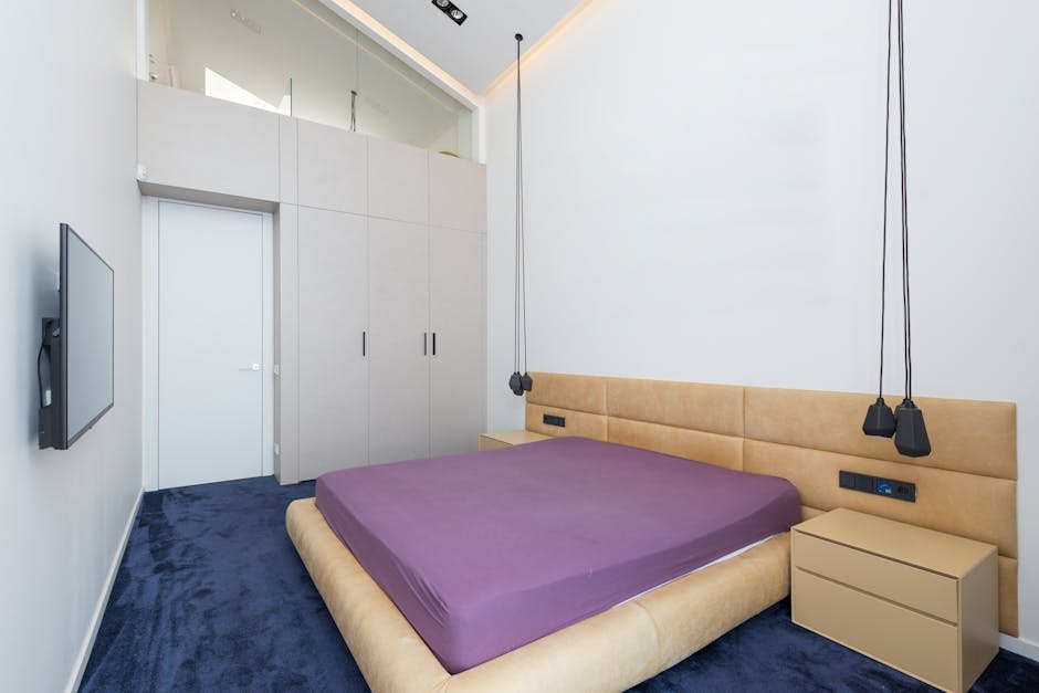 A minimalist bedroom featuring a double bed with a plain white mattress, flanked by two small wooden bedside tables with dark metal legs, positioned against white walls and a white ceiling. To the rig