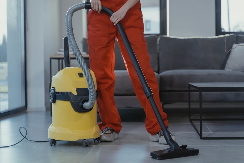 A person dressed in orange overalls is operating a yellow vacuum cleaner with a black hose and an attachment, cleaning a light-colored tiled floor in a modern living room. The room features a dark gra