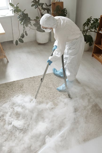 A professional cleaner in full protective gear, including a white suit, gloves, mask, and shoe covers, is performing deep cleaning or sanitisation on a beige carpet in a bright, modern living room. The room features large windows allowing natural light to illuminate light-colored walls, a tall potted plant, and minimal furniture, creating an inviting atmosphere. The cleaner is using a large, adjustable metal wand with a cleaning attachment to thoroughly treat the carpet, which appears to be in the process of being freshened or disinfected. Visible cleaning foam or vapour enhances the impression of intensive surface cleaning typical of services offered by Carpet Cleaning Harringay, focusing on hygiene and maintenance in a domestic setting, aligned with their focus on surface cleaning and deep cleaning solutions.