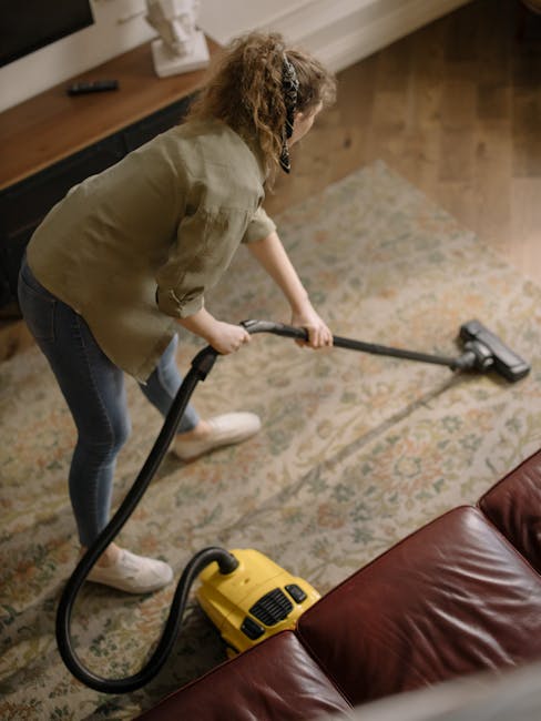 A woman engaged in deep cleaning a patterned area rug in a living room, using a yellow and black vacuum cleaner with a hose attachment. She is wearing casual clothing, including a beige jacket and jeans, and has curly hair. The room features wooden flooring, a leather sofa, and a wooden sideboard with a white decorative object, under warm ambient lighting. The scene depicts a thorough surface cleaning process as part of domestic sanitisation services offered by Carpet Cleaning Harringay, tailored to maintain hygiene and cleanliness in residential settings.