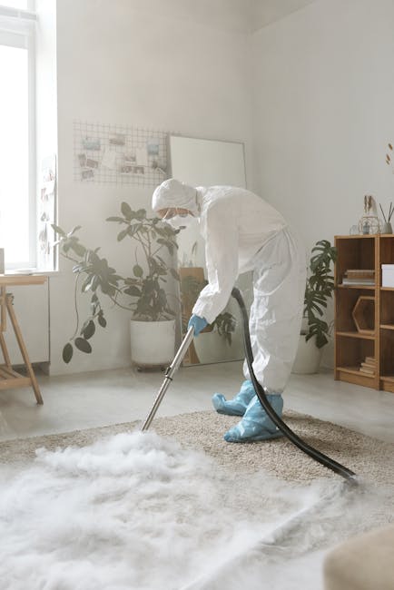 A professional cleaner in full protective gear, including a white suit, gloves, mask, and shoe covers, is performing deep cleaning or sanitisation on a beige carpet in a bright, modern living room. The room features large windows allowing natural light to illuminate light-colored walls, a tall potted plant, and minimal furniture, creating an inviting atmosphere. The cleaner is using a large, adjustable metal wand with a cleaning attachment to thoroughly treat the carpet, which appears to be in the process of being freshened or disinfected. Visible cleaning foam or vapour enhances the impression of intensive surface cleaning typical of services offered by Carpet Cleaning Harringay, focusing on hygiene and maintenance in a domestic setting, aligned with their focus on surface cleaning and deep cleaning solutions.