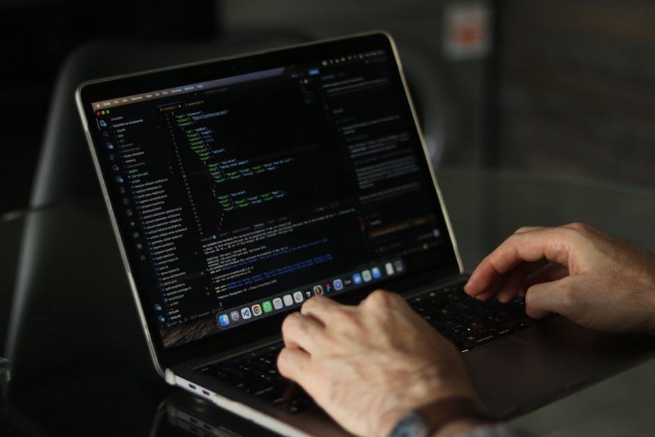 Close-up of a person's hands typing on a laptop keyboard placed on a dark table; the laptop screen displays a dark-themed code editor with lines of programming code. The room has subdued lighting, hig